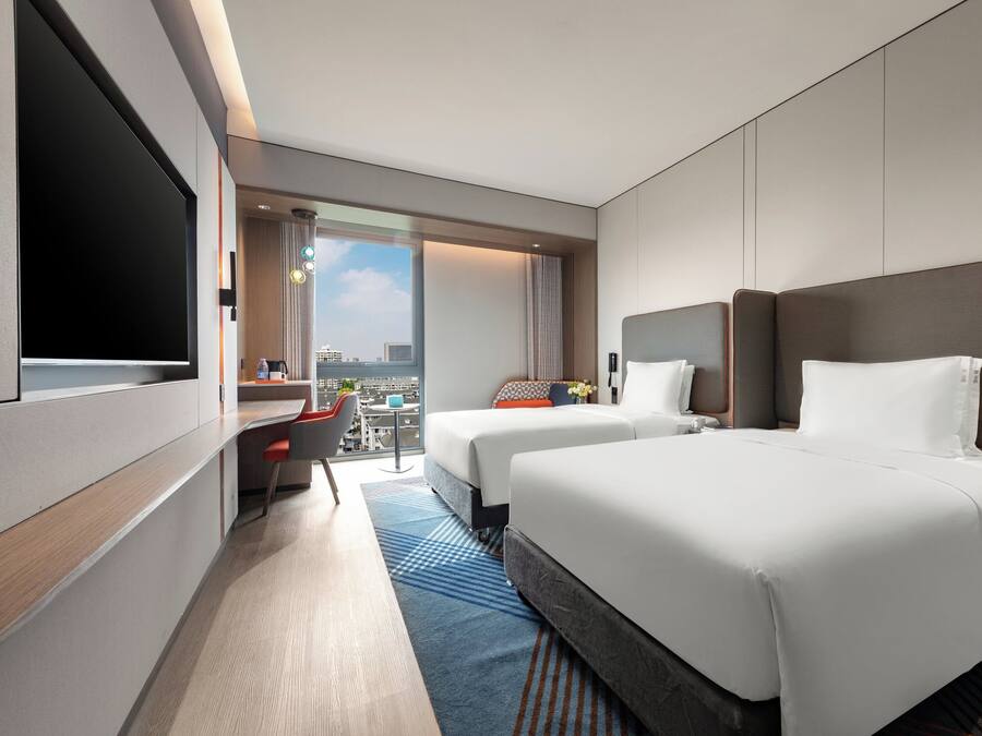 A hotel room with two beds, a desk, a TV, and a view of the city.