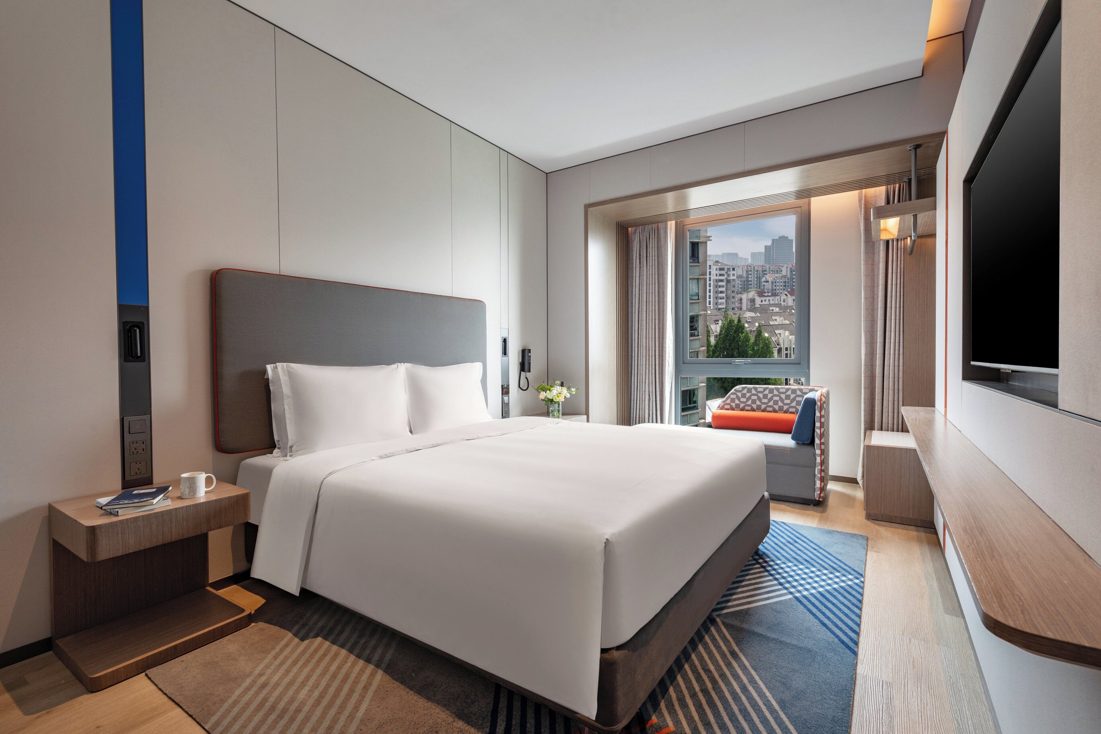 A hotel room with a large bed, a bedside table with a cup and book, a TV, and a view of the city through the window.