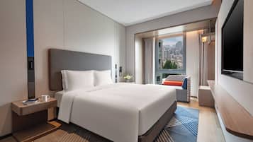 A hotel room with a large bed, a bedside table with a cup and book, a TV, and a view of the city through the window.