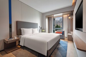 Una camera d'albergo con un letto grande, un comodino con una tazza e un libro, una TV e vista sulla città dalla finestra.