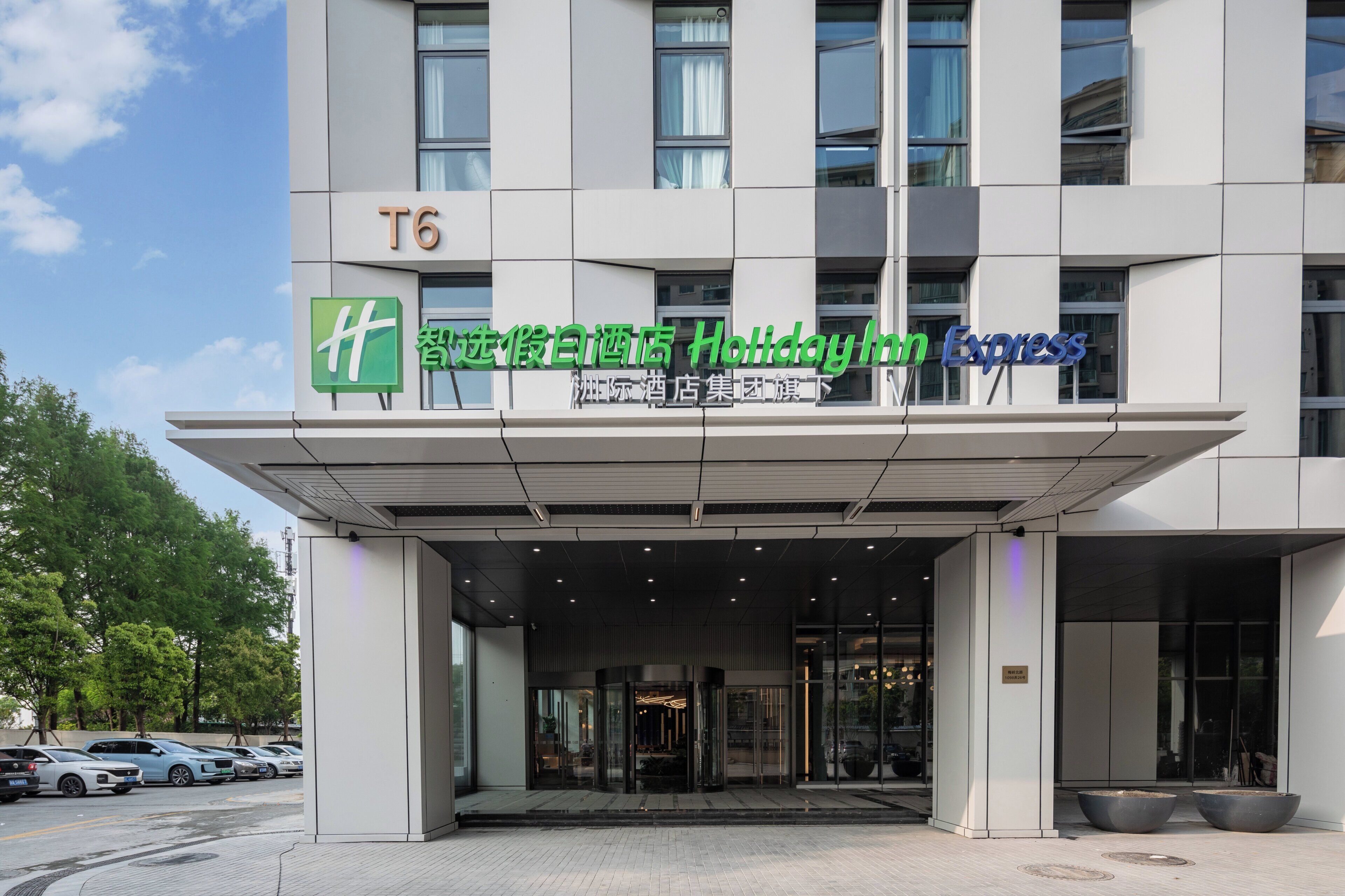A hotel entrance with a clear sky, a parking lot with cars, and a sign indicating it's part of the Holiday Inn Express chain.
