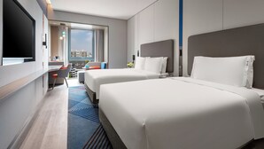 A hotel room with two beds, a TV, a desk with a chair, a window with a view of buildings, and a balcony.