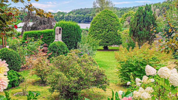 Un jardín bien cuidado con diversos arbustos, una estructura de piedra y una vista despejada de un lago al fondo.
