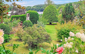 A well-maintained garden with various shrubs and flowers, a stone structure, and a view of a lake in the background.