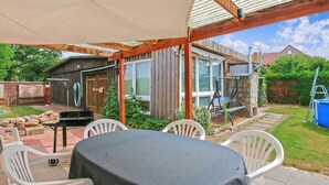 En overdækket terrasse med et bord og stole, grill og en træbygning med ståltag.