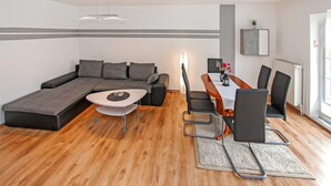Ein modernes Wohnzimmer mit einem Ecksofa, einem Esstisch mit Stühlen, einem kleinen runden Tisch und einem einzelnen Sessel sowie einer Stehlampe.