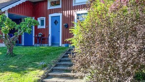 A red wooden house with a blue door and window, a small garden with a tree and a bush, and a stone staircase leading to the front door.