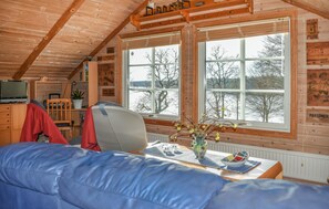 Une chambre confortable de style cabane, avec un plafond en bois, une grande fenêtre, un téléviseur, un fauteuil, une table avec un vase et un canapé bleu.