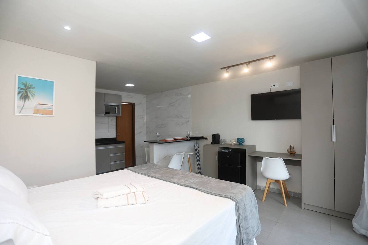 A hotel room with a bed, a desk with a chair, a wardrobe, a TV mounted on the wall, and a kitchen area in the background.