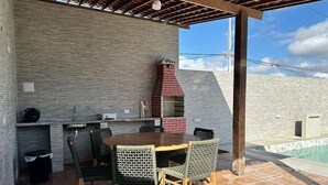 An outdoor patio with a brick wall, a wooden table with chairs, a grill, and a pool. - Solaris Serrambi Flats (Ipojuca)