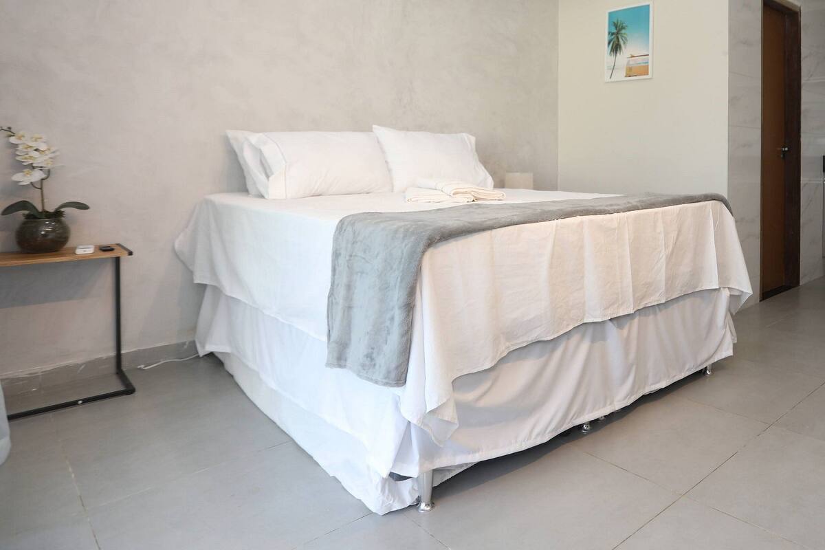 A bedroom with a bed, white walls, a ceiling-mounted air conditioner, a small table with a vase, and a door.