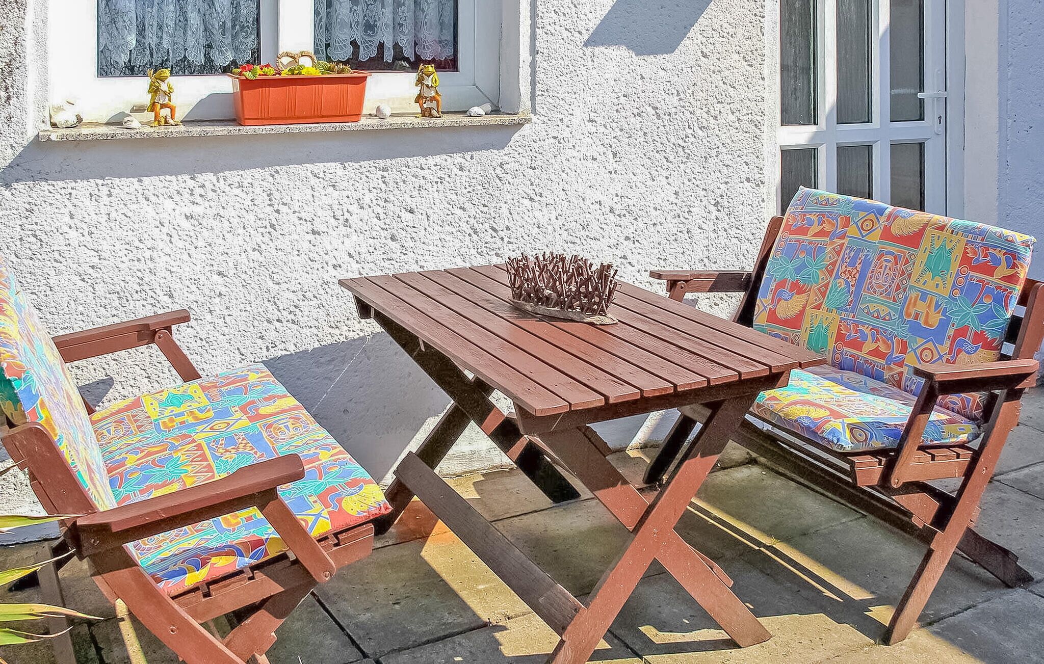 Uma mesa de madeira e duas cadeiras com almofadas coloridas, colocadas em frente a um edifício com fachada branca e uma janela com cortinas de renda.