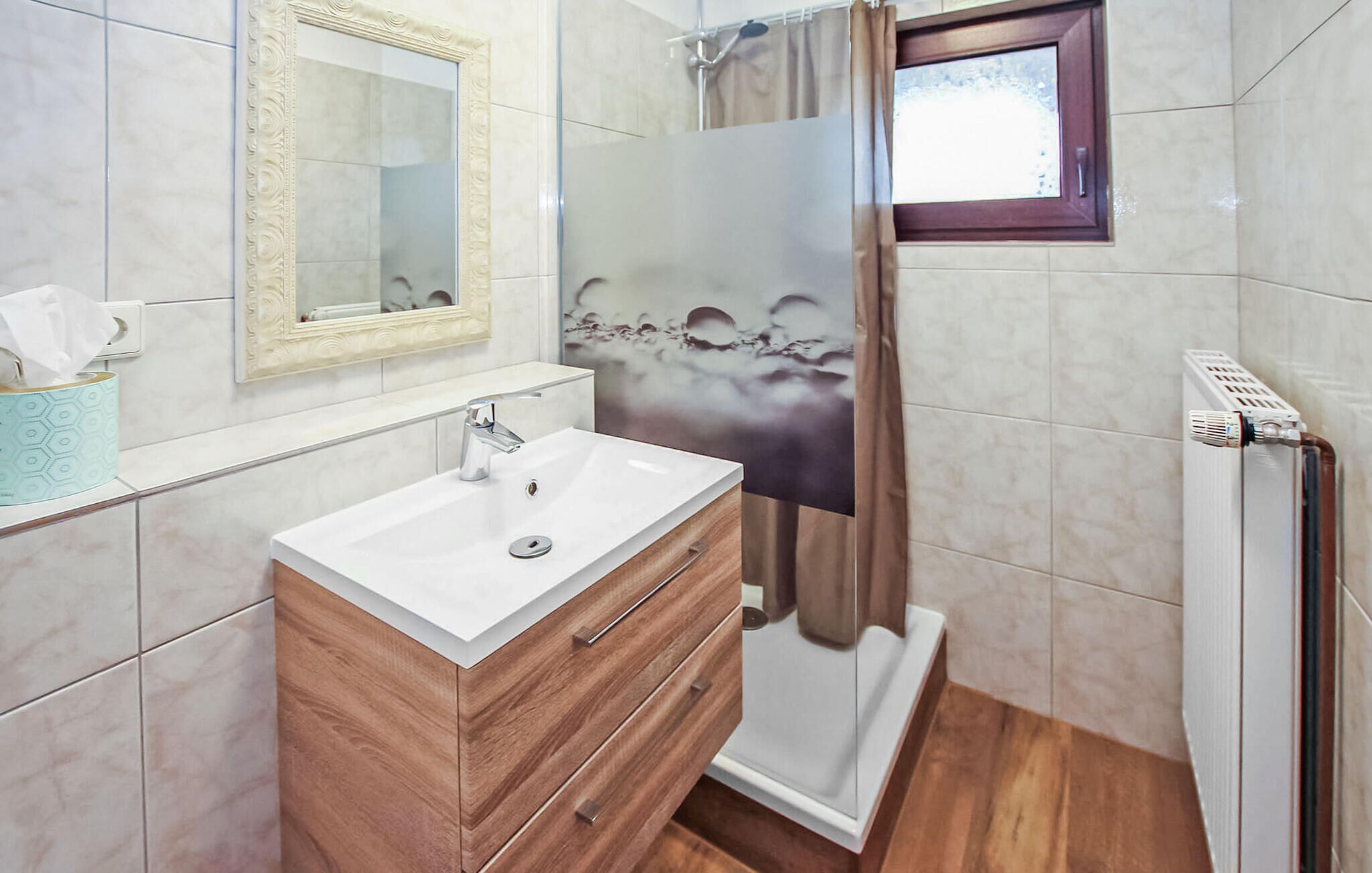 Ein Badezimmer mit weißem Waschbecken, Holz-Waschtisch, einem Spiegel, einer Dusche mit Glastür und einem Fenster.