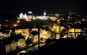 Une vue nocturne d’un château historique, avec ses tours illuminées et son imposant dôme, entouré d’une ville dont les bâtiments sont éclairés, ainsi que d’un pont.
