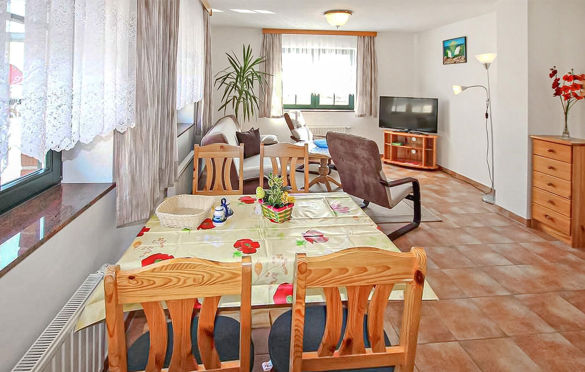 Un comedor con una mesa lista para cuatro personas, rodeada de sillas de madera. Un televisor está montado en la pared y hay un mueble con cajones a la derecha.
