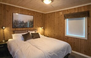 Un dormitorio con un revestimiento de madera en la pared, una cama con ropa blanca, dos lámparas de noche, un cuadro enmarcado en la pared y una ventana con persiana.