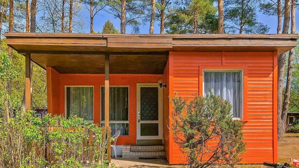 Une petite cabane à un seul étage, à l’extérieur rouge, avec un porche doté d’un banc et offrant une vue sur la forêt environnante.
