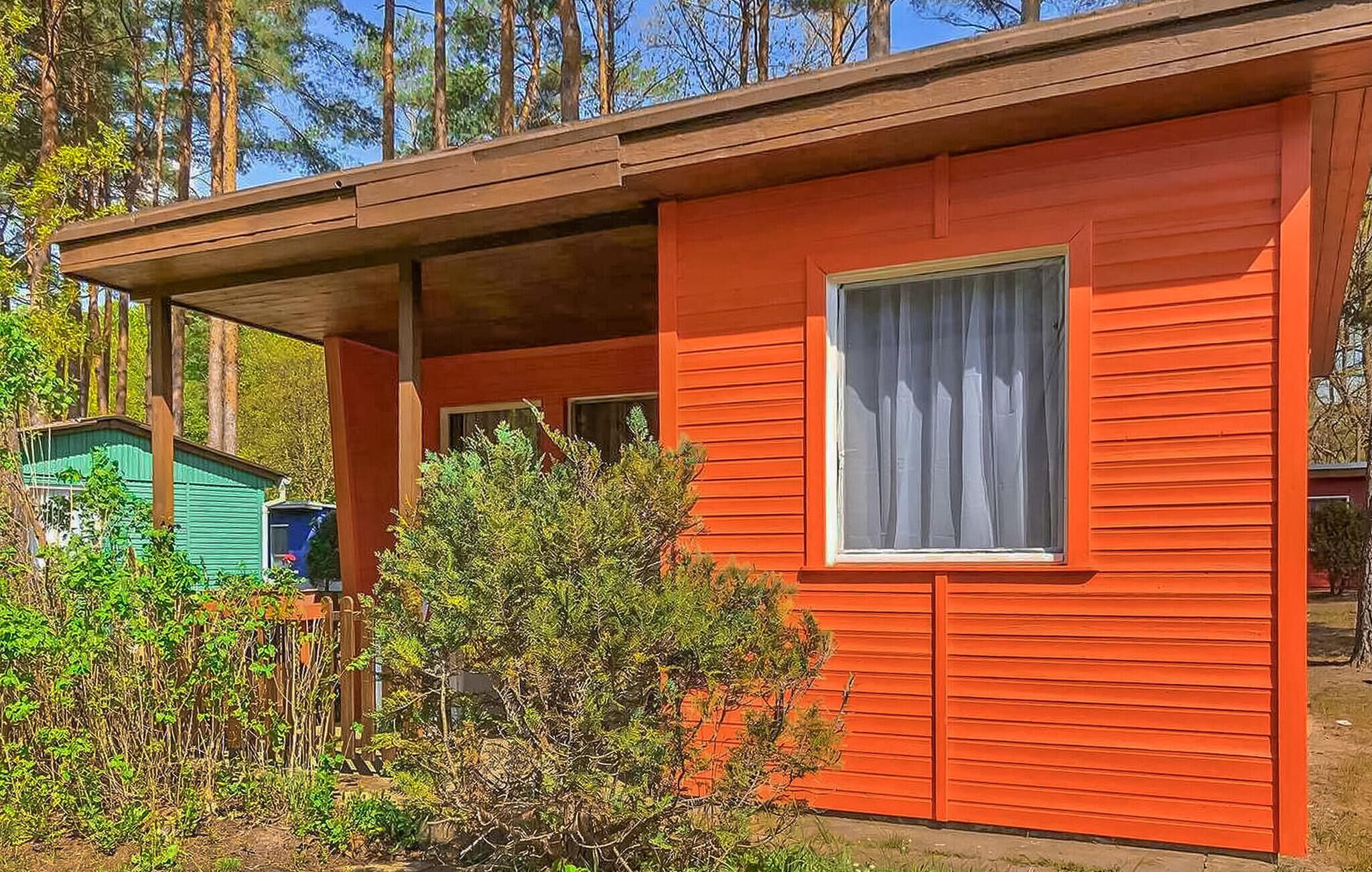 A red cabin with a covered porch, a window with white curtains, and a green shed in the background.