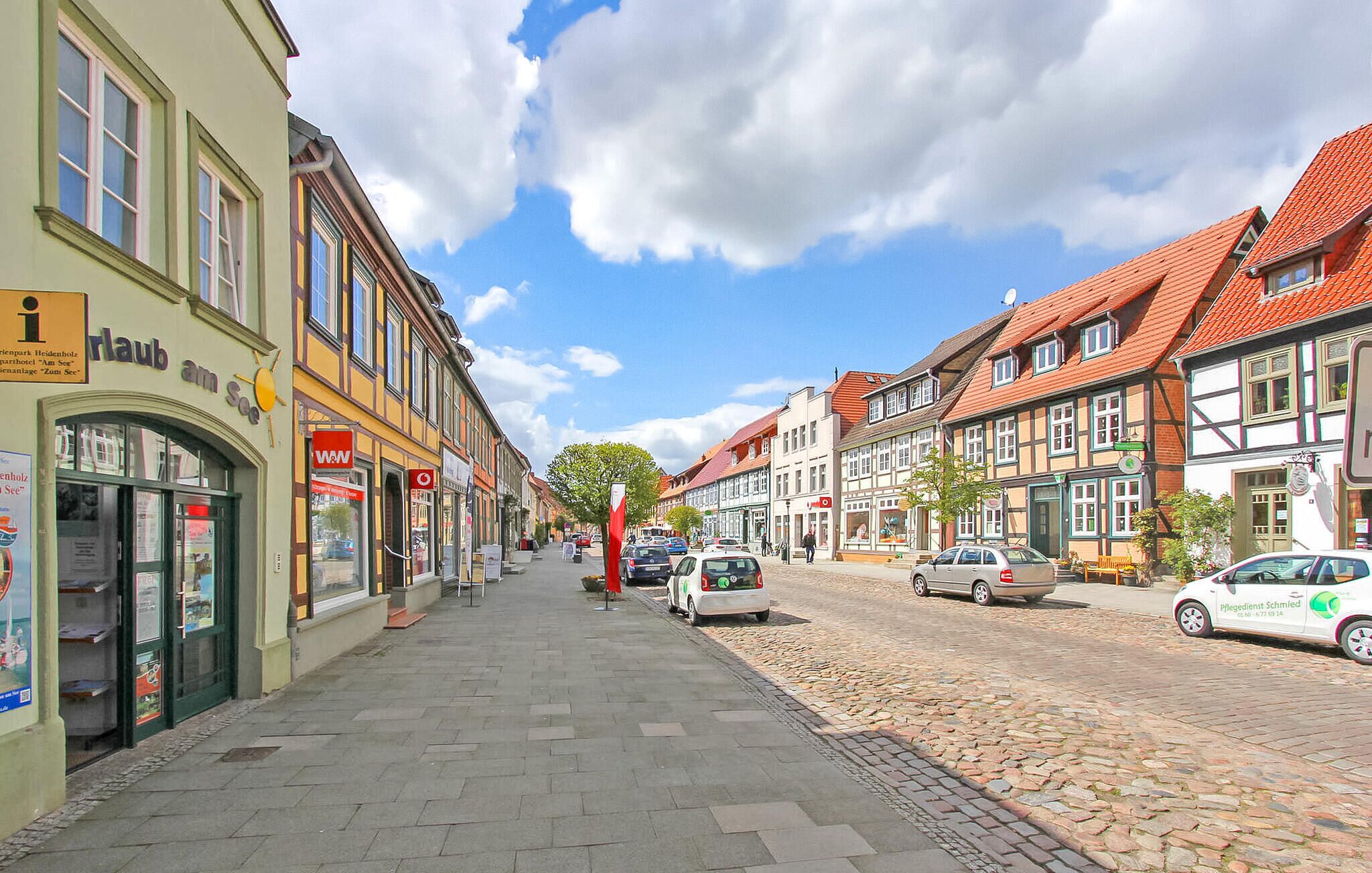 A cobblestone street lined with traditional European buildings, a clear sky, and parked cars.