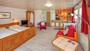 Ein Zimmer mit einem Bett, einem roten Sessel, einem kleinen Tisch mit einer Zeitung, einem Fernseher, einem Spiegel, einer Küchenzeile mit Schränken und einer Mikrowelle sowie einem roten Vorhang.