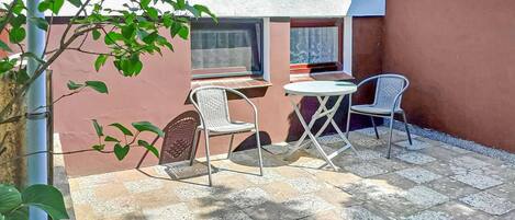 An outdoor patio with two wicker chairs and a small round table, a window, and a plant.