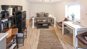 Un espace de vie compact comprenant un coin cuisine, un lit avec une tête de lit, un coin repas et une fenêtre agrémentée d’une plante.
