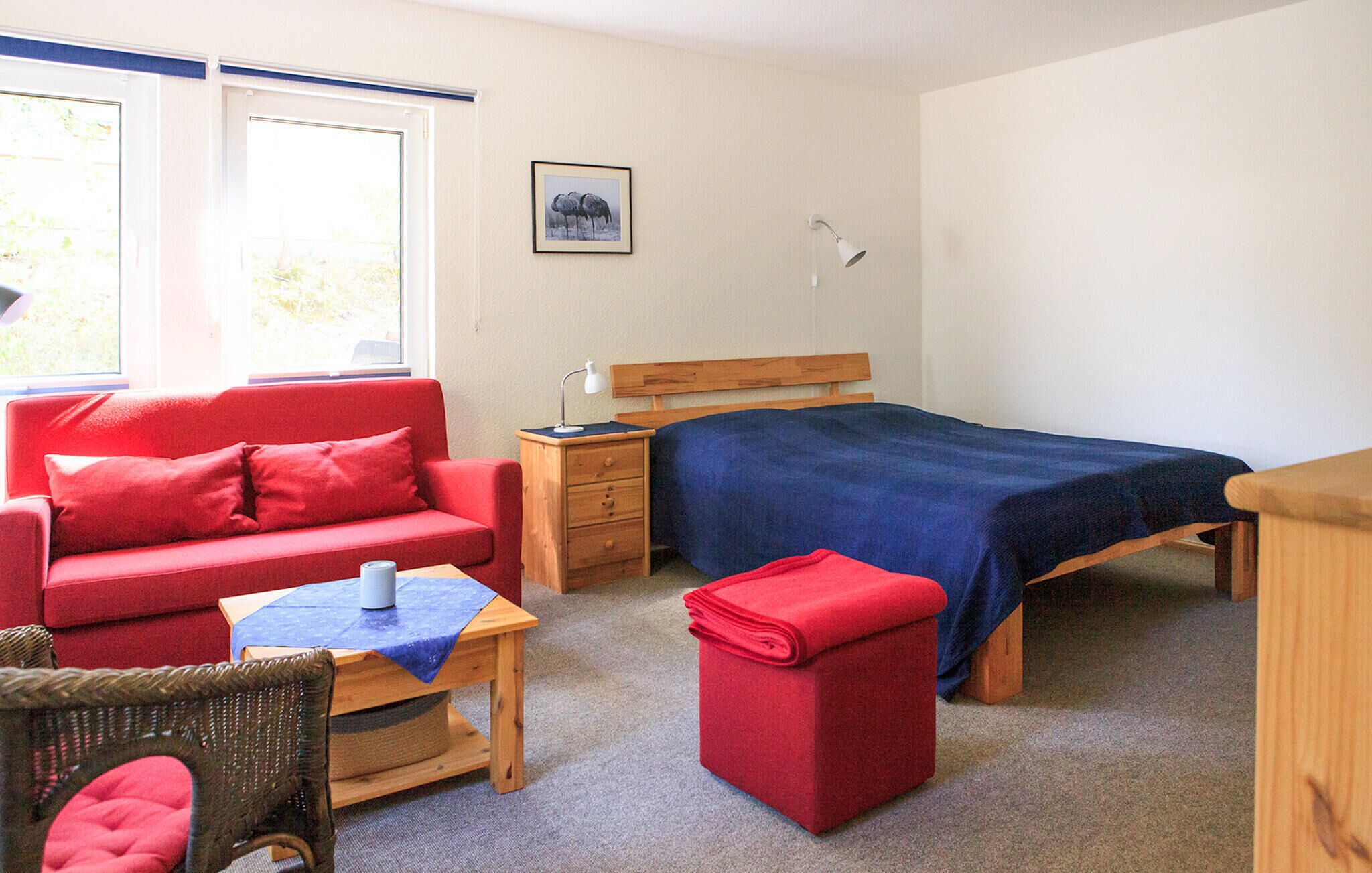 Een kamer met een rode bank, een houten bed met een blauwe deken, een houten nachtkastje, een rieten stoel en een kleine tafel met een blauwe tafelkleed.