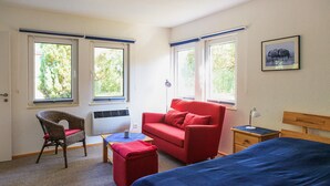 A room with a bed, a red sofa, a wicker chair, a small table, and two windows. - Ferienwohnung Rheinsberg (Rheinsberg OT Kagar)