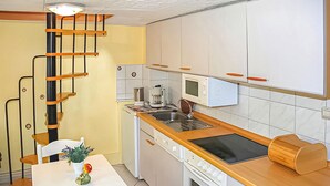 Eine kleine Küche mit integriertem Herd, Mikrowelle und Kaffeemaschine. Ein Holztisch ist mit einem Blumentopf und einem Stuhl bestückt.