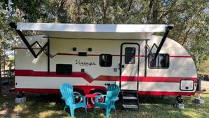 A vintage RV with a covered front, two blue chairs, and a small step leading to the door. - Retro Style Camper. SW Ocala-Near WEC-Comfy Bed (Ocala)