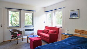 A room with a bed, a red sofa, a wicker chair, a small table, and two windows with blue blinds.
