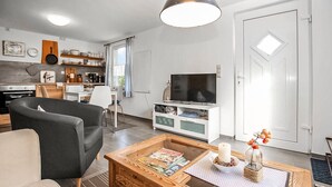 Een moderne woonkamer met een bank, salontafel en televisie. Achtergrond toont een keuken met een eethoek.
