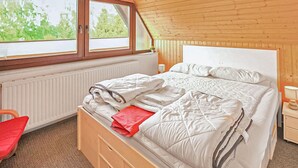 Une chambre avec un plafond en bois, un lit recouvert de linge de lit blanc, une chaise rouge et une fenêtre donnant sur des arbres.
