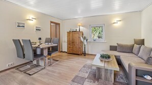 Ein modernes Wohnzimmer mit einem Holzesstisch, zwei Stühlen, einem Sofa und einem Fenster mit Blick auf Grünflächen.