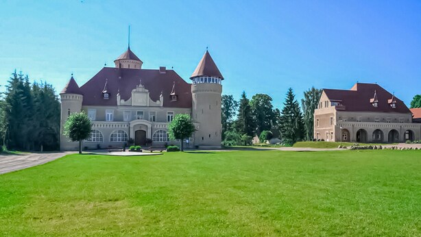 Ein großes, historisches Gebäude mit einem zentralen Turm und zwei kleineren Türmen, umgeben von einer gepflegten Rasenfläche und Bäumen.