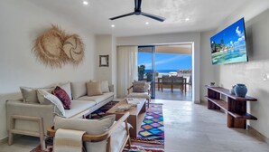A spacious living room with a sofa, chairs, a coffee table, and a TV. There is a ceiling fan, a large window with a view of the ocean, and a colorful rug.