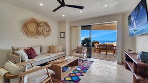 A modern living room with a sofa, coffee table, and chairs, a ceiling fan, and a view of the ocean through a sliding glass door.