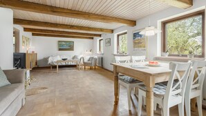 Amplia sala de estar con techo de madera, comedor con mobiliario blanco y vista a zonas verdes a través de ventanas grandes.