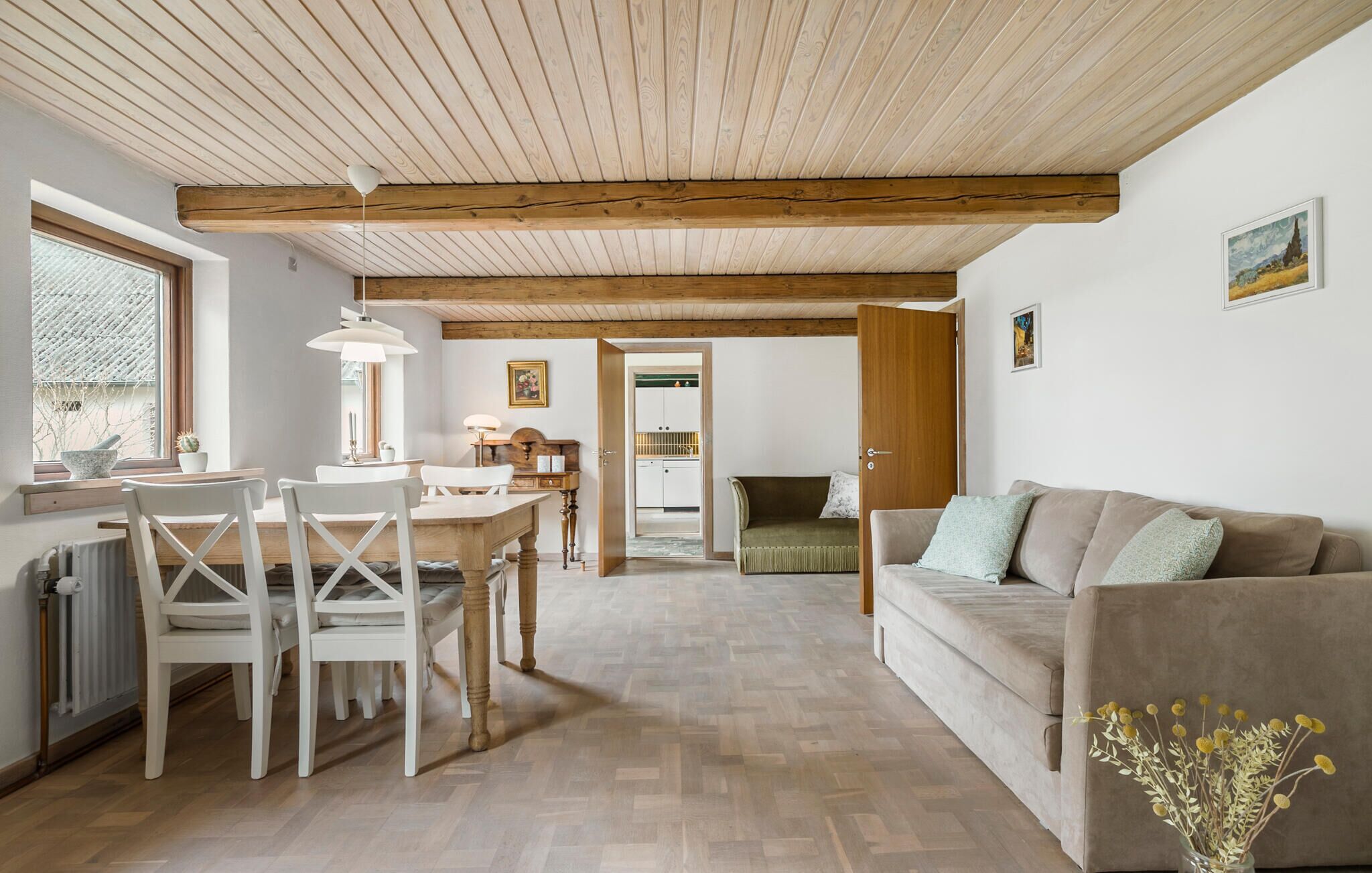 A cozy living room with a wooden ceiling, a dining area with white chairs, a sofa, and a vase with flowers.