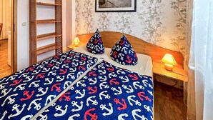 Ett sovrum med en mörkblå sängkappa prydd med ankarmotiv, två kuddar med liknande mönster, en sänggavel i trä, ett nattduksbord med en lampa och en inramad tavla på väggen.