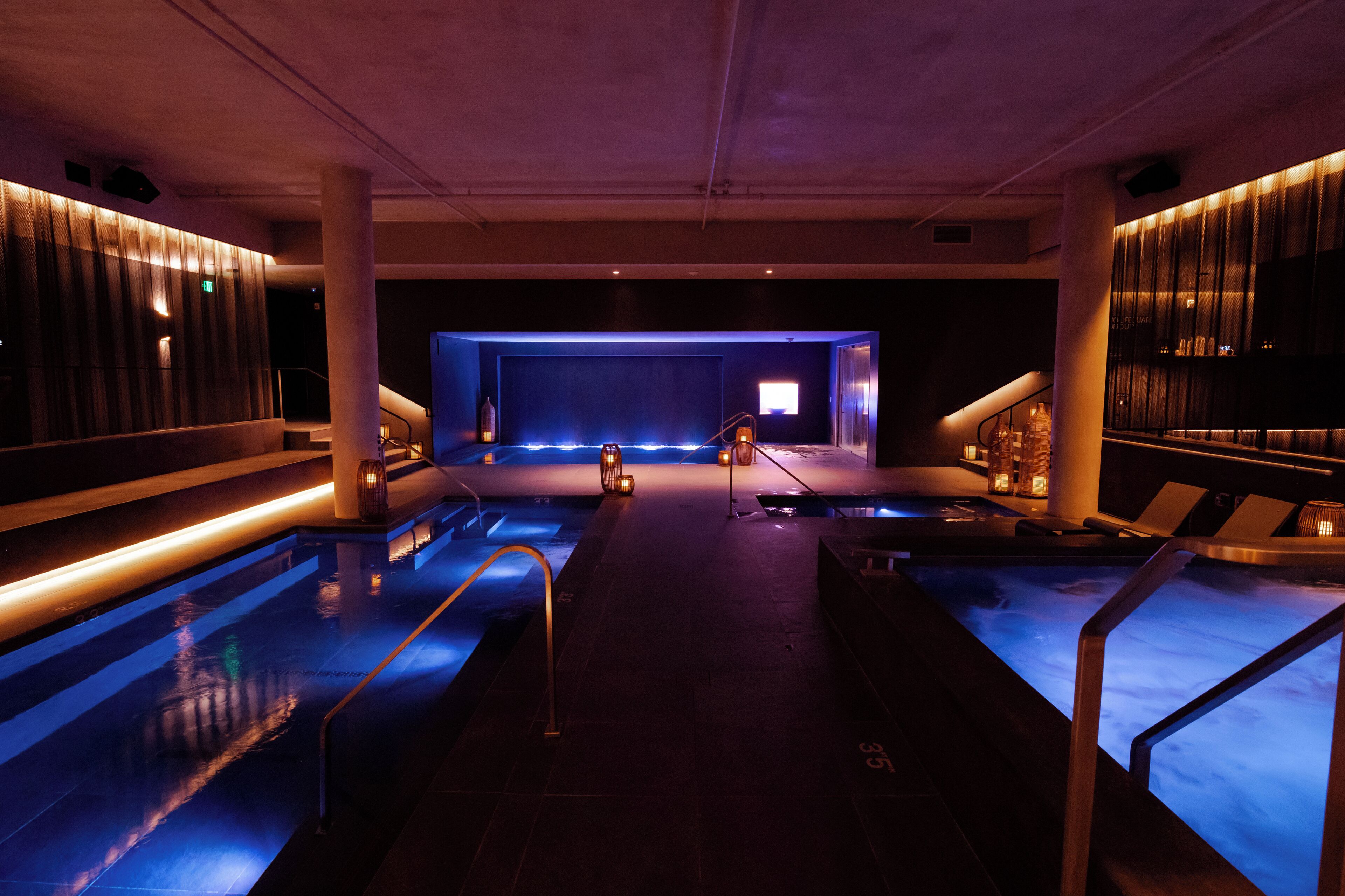 A modern indoor pool area with multiple pools, a central stage, and ambient lighting.
