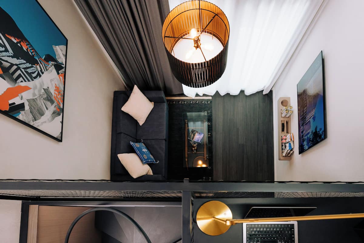 A modern hotel room with a dark-colored sofa, a large abstract painting, a round pendant light, and a laptop on a desk.