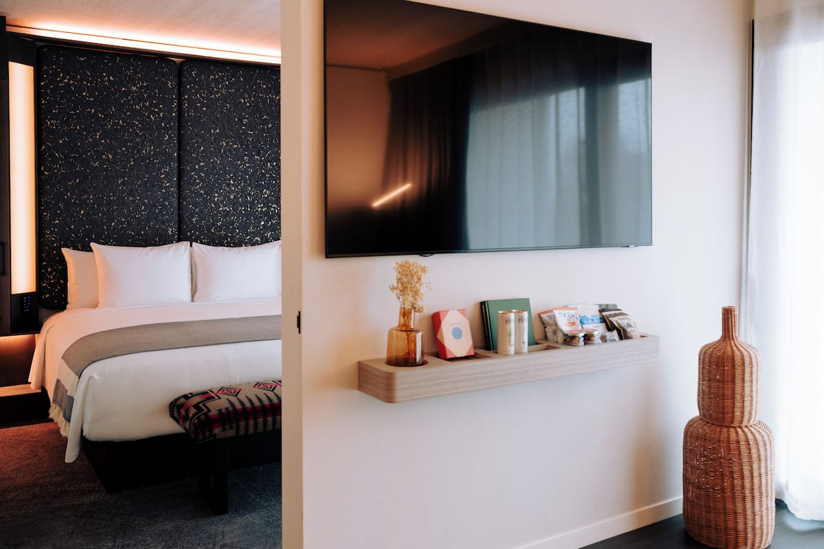 A modern hotel room with a large bed, a mounted TV, a bedside table with books and a vase, and a decorative wicker vase.