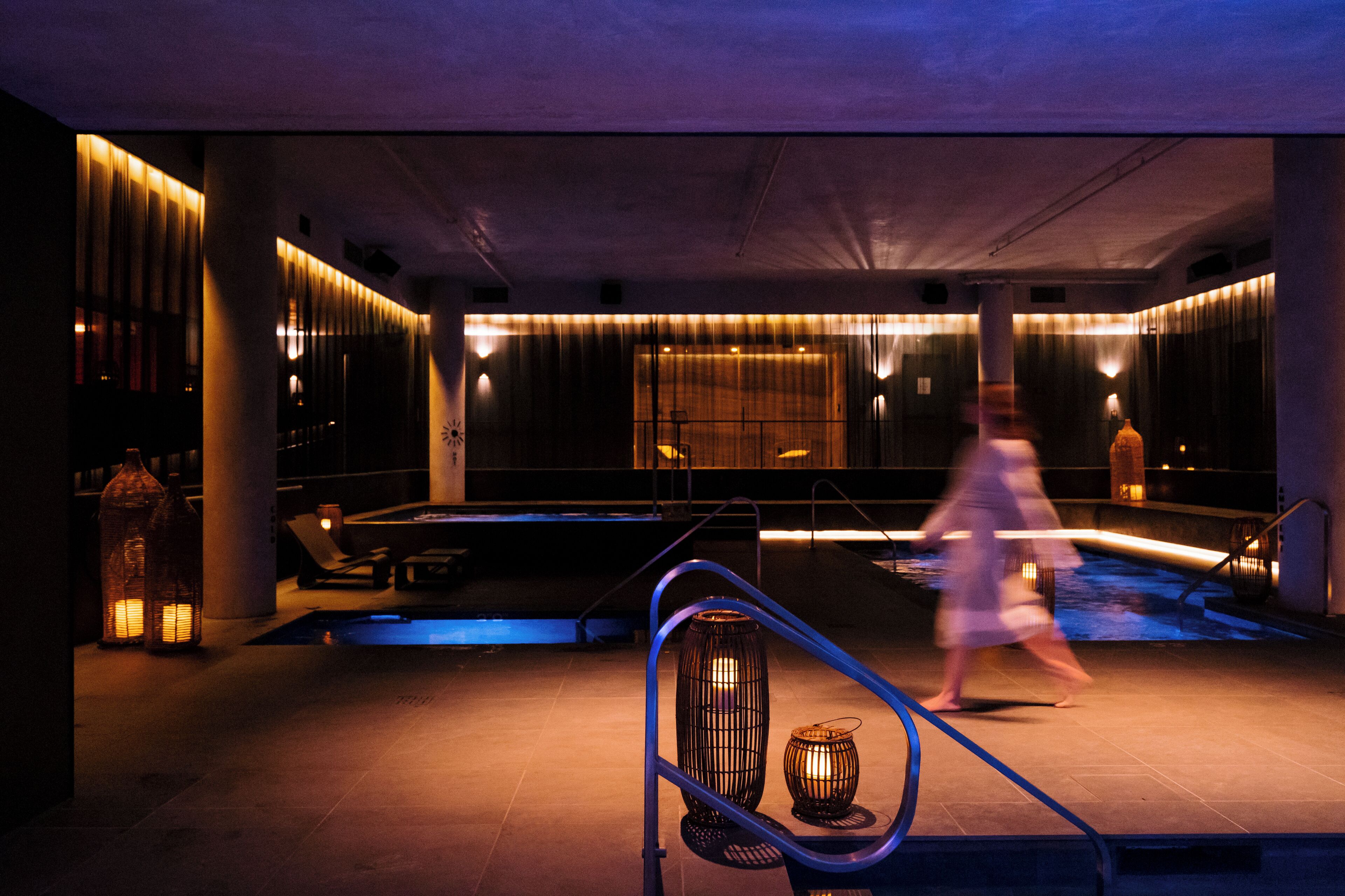 A dimly lit indoor pool area with a modern design, featuring a glass barrier, lounge chairs, and ambient lighting.