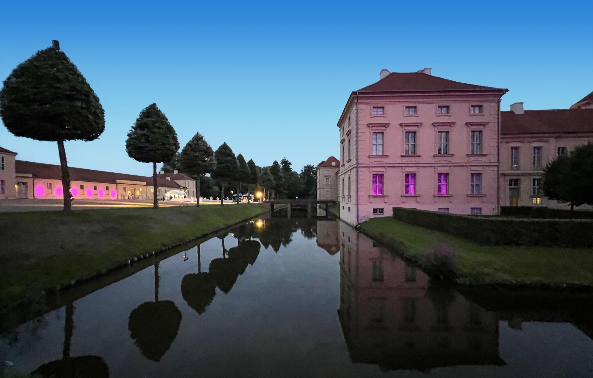 Ein ruhiges Gewässer spiegelt Gebäude wider, darunter ein markantes pinkfarbenes Gebäude mit vielen Fenstern und einem roten Dach.