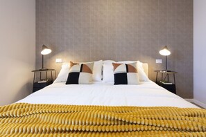 Un lit bien fait, recouvert d’une couverture à motifs, avec deux lampes de chevet et un mur à la texture intéressante.