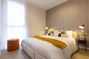 A bedroom with a bed, bedside lamps, a small orange ottoman, and a window with curtains.