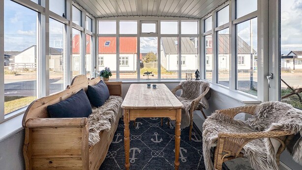 Ein Wintergarten mit einer hölzernen Bank, einem Tisch und Korbstühlen, ausgestattet mit großen Fenstern und Blick auf Häuser.