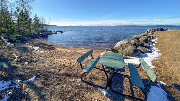 Una playa con una mesa y sillas de picnic, rodeada de rocas y árboles, con vista al agua y la costa distante.