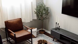 Ein modernes Wohnzimmer mit einem dunkelblauen Sofa, einem braunen Ledersessel, einem Holztisch mit einer dekorativen Schale, einem kleinen Beistelltisch, einer Topfpflanze und einer TV-Konsole mit Kerzen.